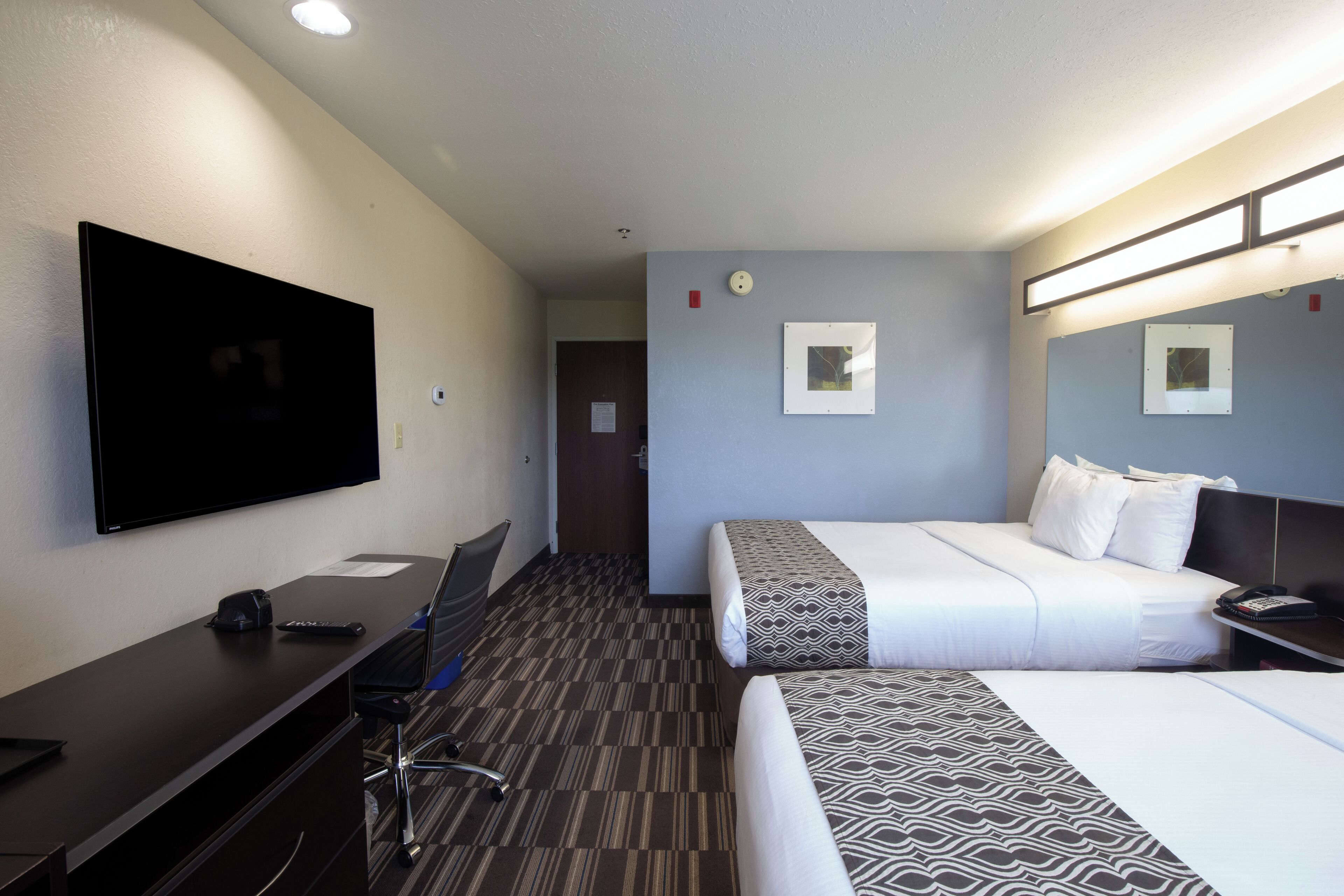 Habitación de hotel con televisión de pantalla plana, escritorio, silla, cama con ropa de cama estampada y mesita de noche con teléfono.