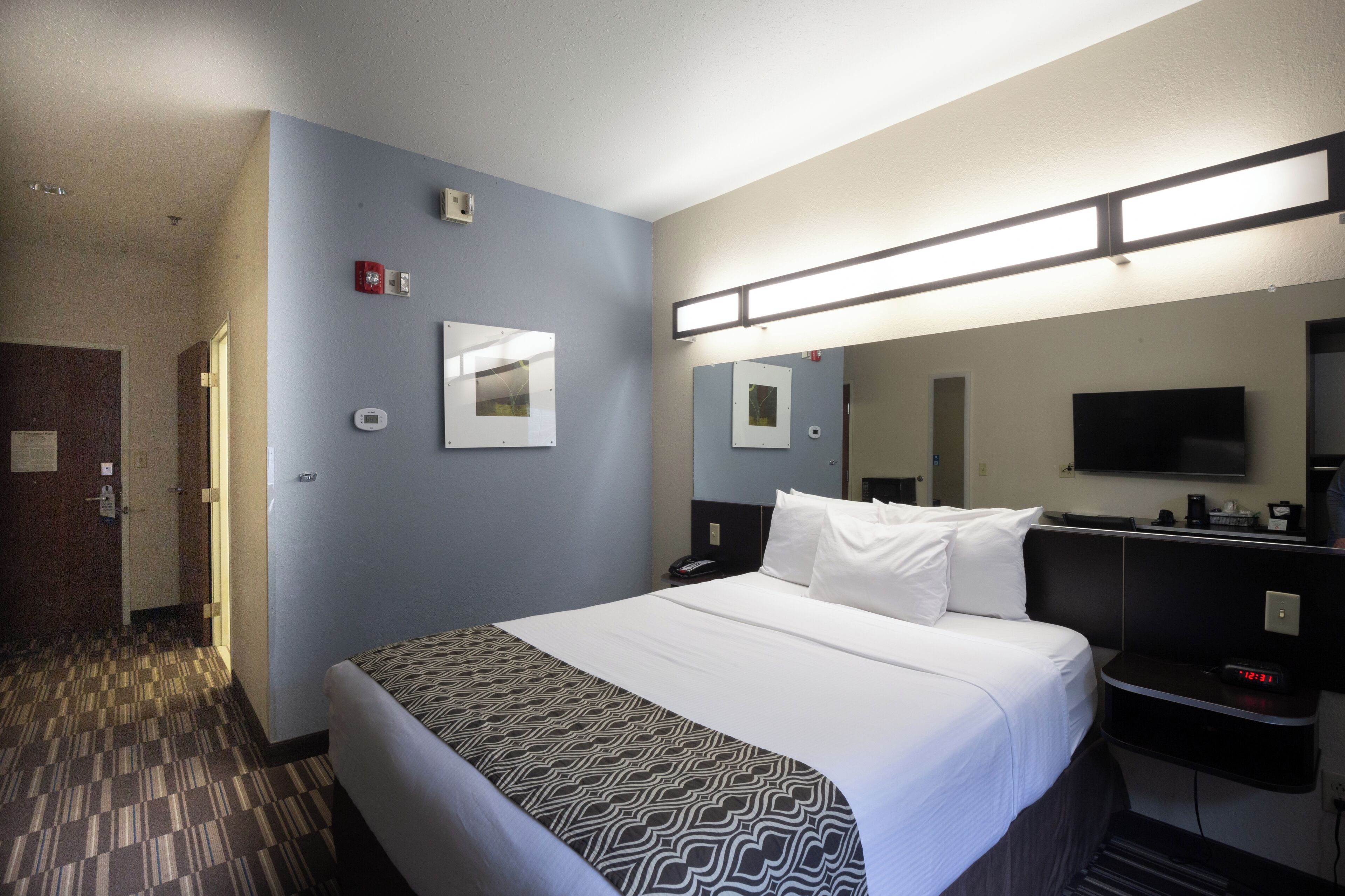 A hotel room with a large bed, a bedside table with a lamp, a flat-screen TV, and a door leading to another room.