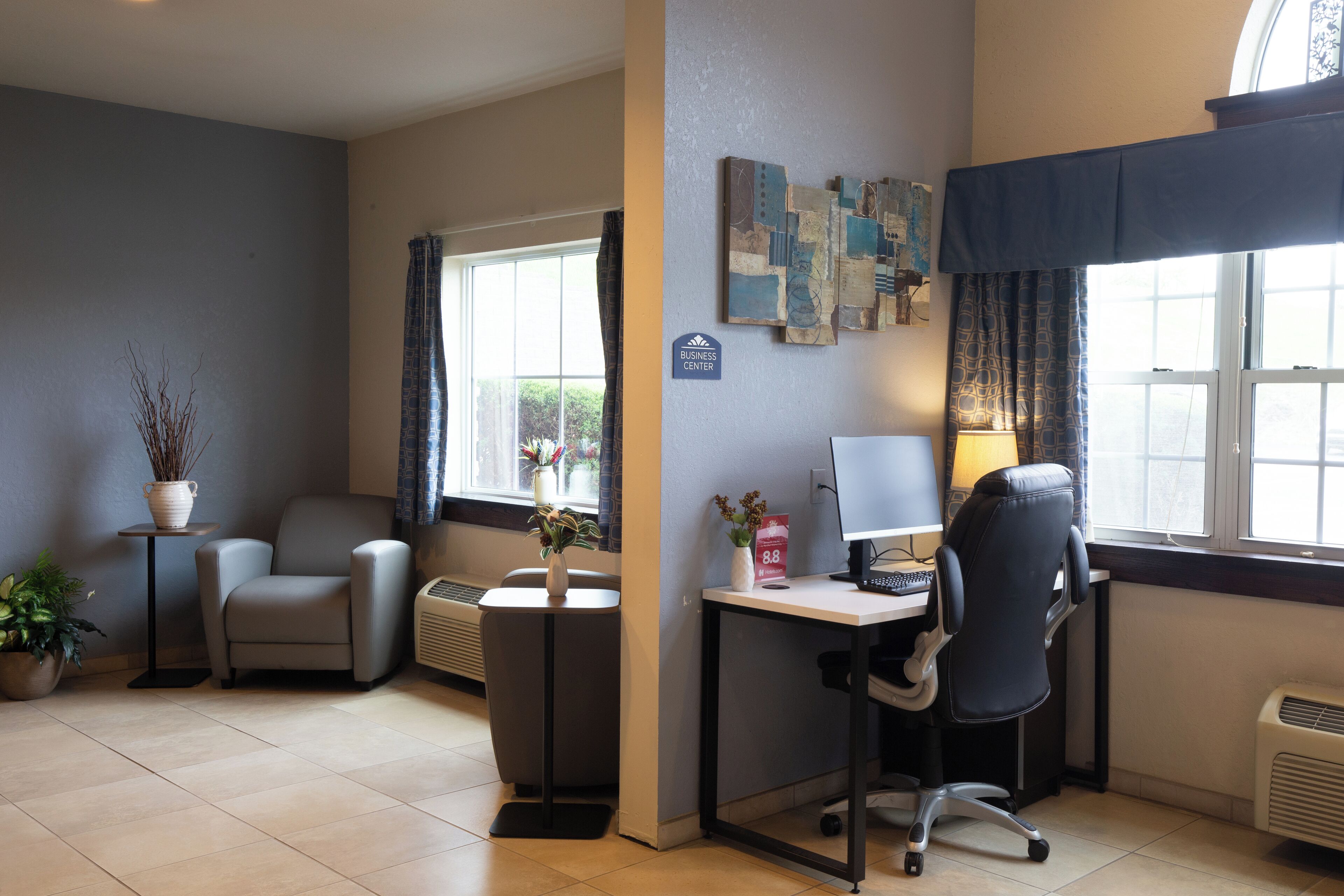A modern office space with a desk, computer, and two chairs. There are potted plants and a window with curtains.