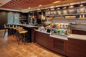 Zona de bar con una barra larga, taburetes, una vitrina refrigerada con bebidas y un panel con la carta sobre la barra.
