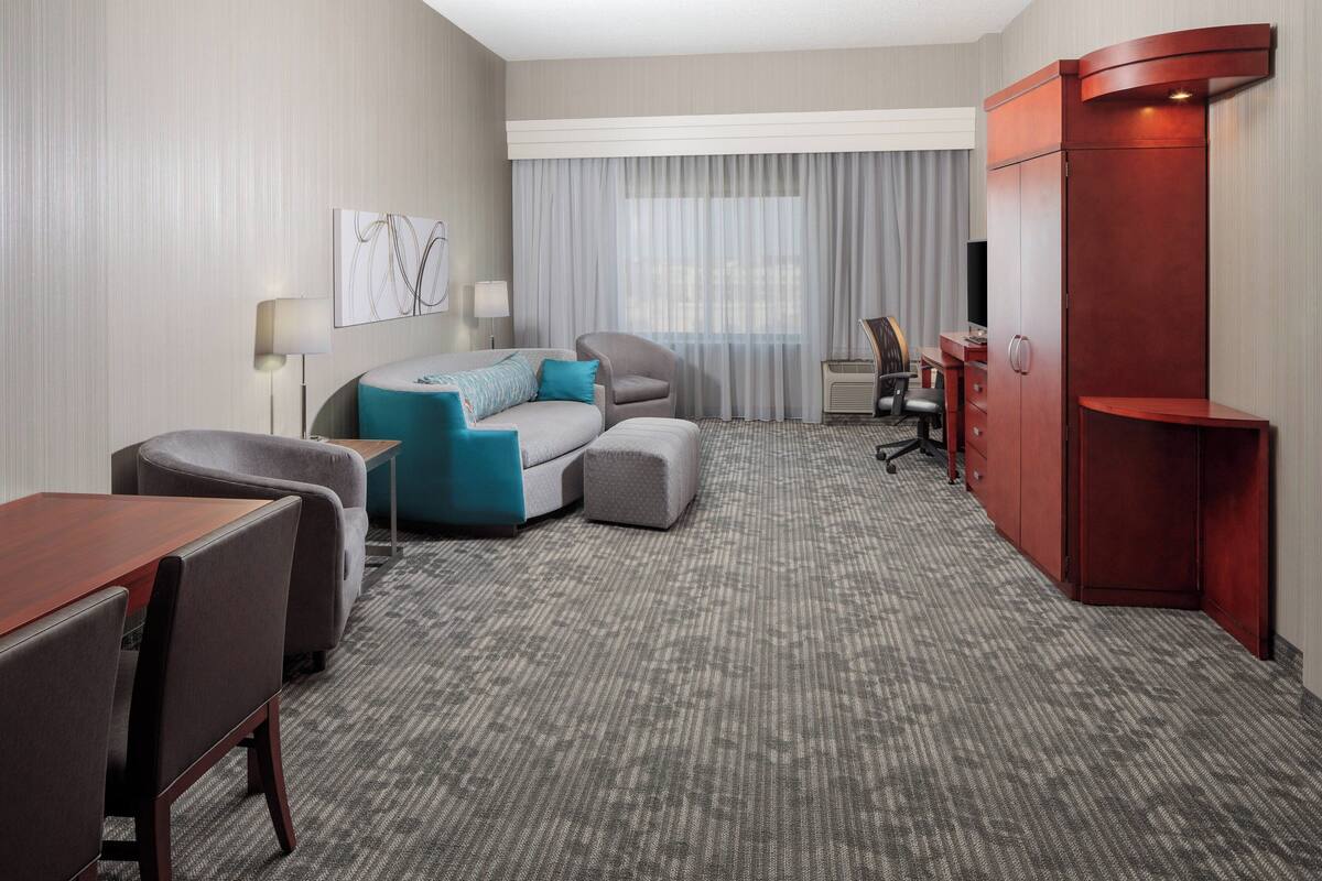 Une chambre d’hôtel avec un tapis gris, un canapé gris agrémenté de coussins bleus, un bureau en bois, une armoire rouge et un téléviseur.