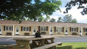 A motel with a row of rooms, a picnic table, and a parking lot. - All Seasons Inn Corp (Oswego)