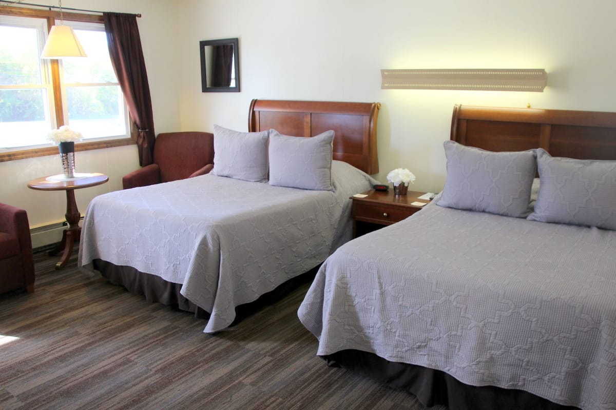 A hotel room with two beds, a sitting area with a chair, a small table with a lamp, and a window with curtains.