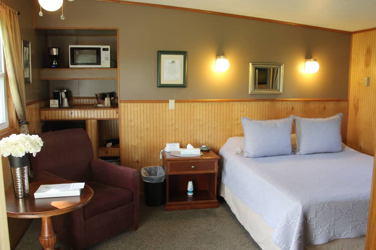 A hotel room with a bed, a desk with a chair, a microwave, a trash can, and a small table with a vase of flowers.