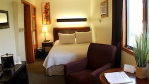A hotel room with a bed, a chair, a desk with a lamp, a window with curtains, and a plant.