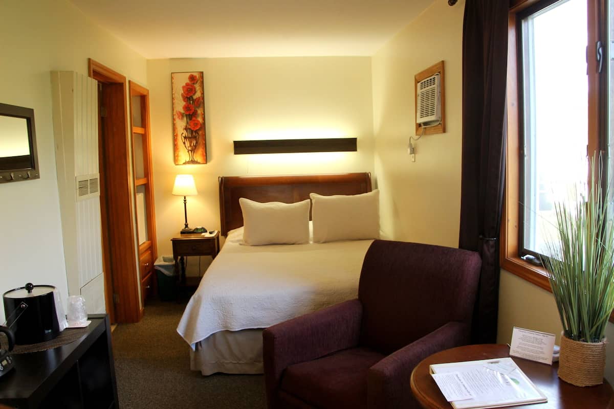 A hotel room with a bed, a chair, a desk with a lamp, a window with curtains, and a plant.