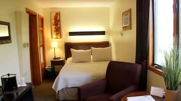 Habitación de hotel con cama, una silla, escritorio con lámpara, ventana con cortinas y planta.