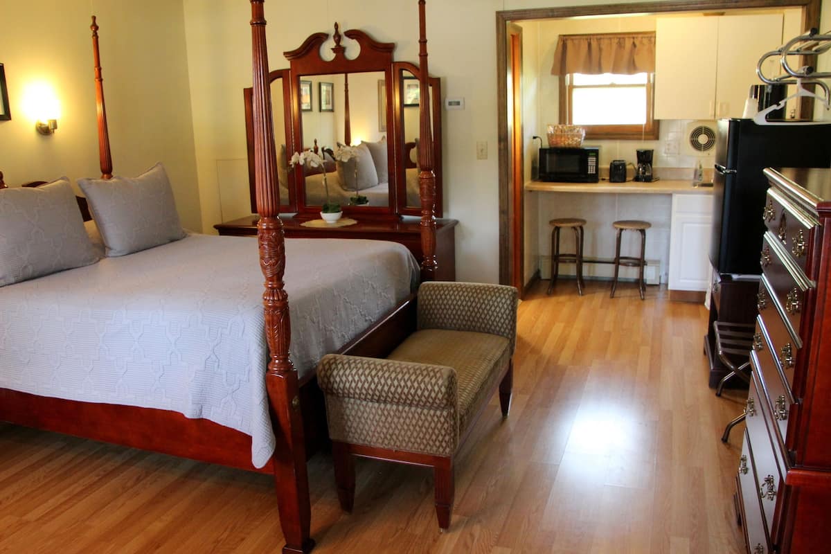 A bedroom with a four-poster bed, a sitting area with a chair, a small kitchen area with a microwave, and a bar stool.