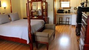 A bedroom with a four-poster bed, a sitting area with a chair, a small kitchen area with a microwave, and a bar stool.