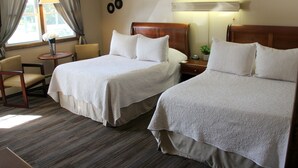 A hotel room with two beds, a small table with a lamp, a chair, and a window with curtains. - All Seasons Inn Corp (Oswego)