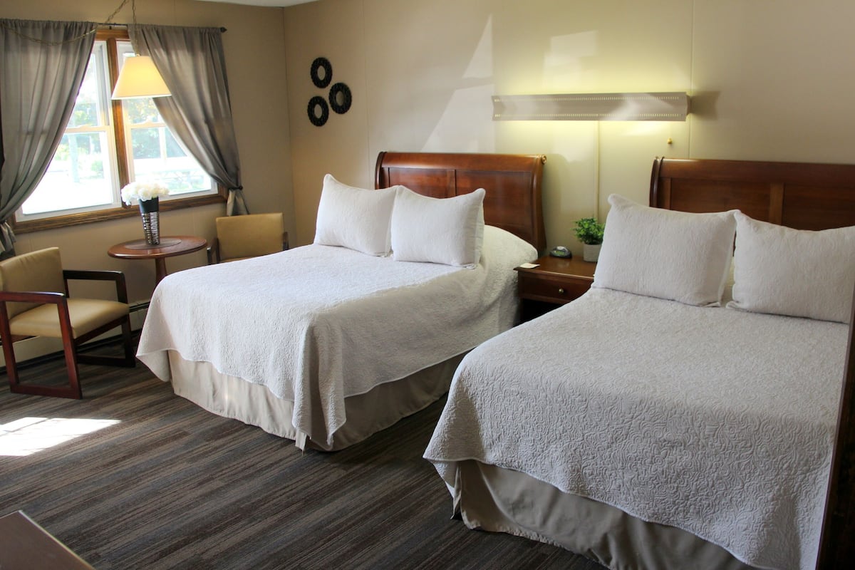A hotel room with two beds, a small table with a lamp, a chair, and a window with curtains.