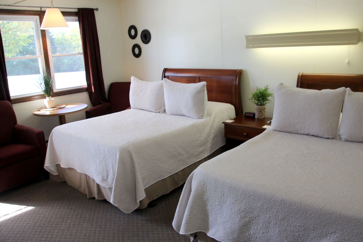 A hotel room with two beds, a sitting area with a chair, a small table, and a window with curtains.