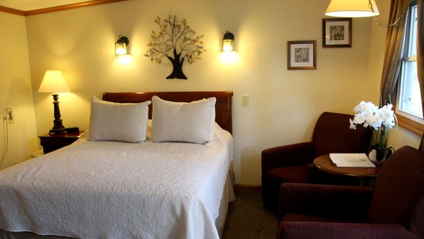 A hotel room with a large bed, two bedside lamps, a nightstand, a chair, a small table, and a vase with flowers. - All Seasons Inn Corp (Oswego)