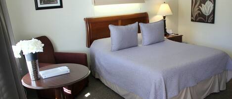 Una habitación de hotel con una cama, una mesita de noche con lámpara, una silla, una pequeña mesa redonda con un florero y un cuadro en la pared.