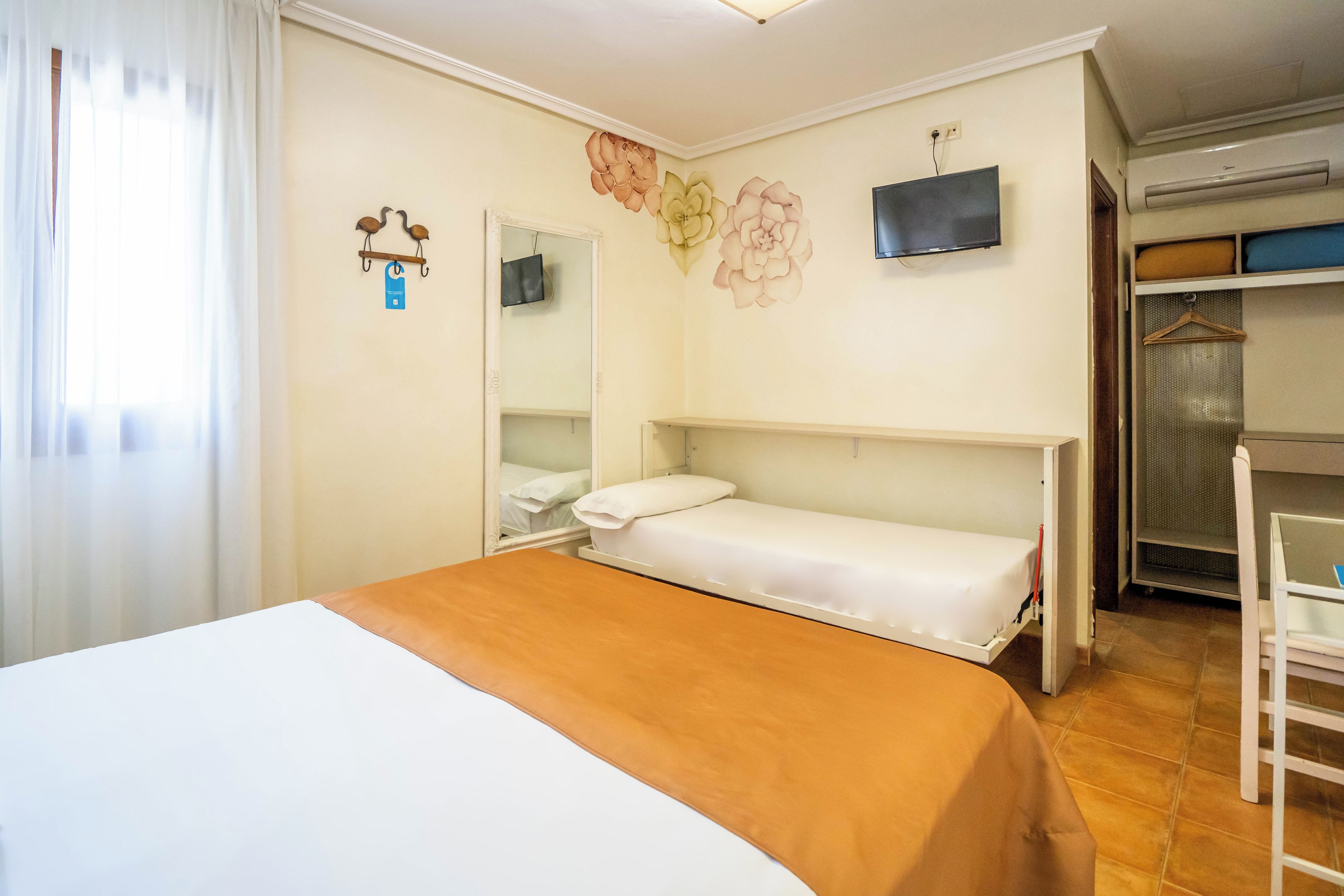 Ein Einzelbett mit weißer und orangefarbener Bettdecke, ein Nachttisch, ein Spiegel, ein an der Wand befestigtes Fernsehgerät und ein Kleiderschrank mit Platz zum Aufhängen von Kleidung.
