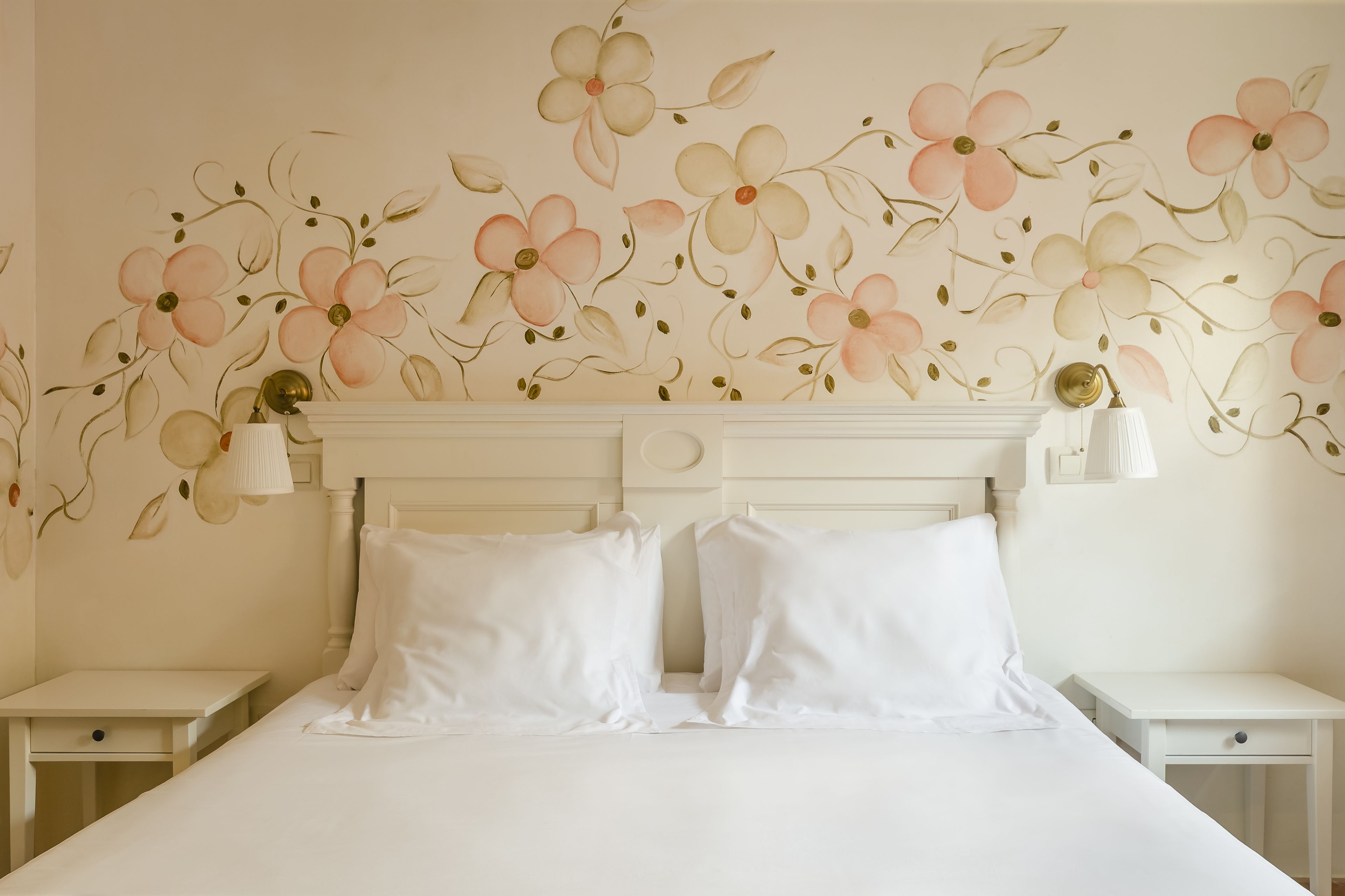 Una cama bien tendida con sábanas blancas, un mural floral y lámparas de noche sobre mesitas blancas.