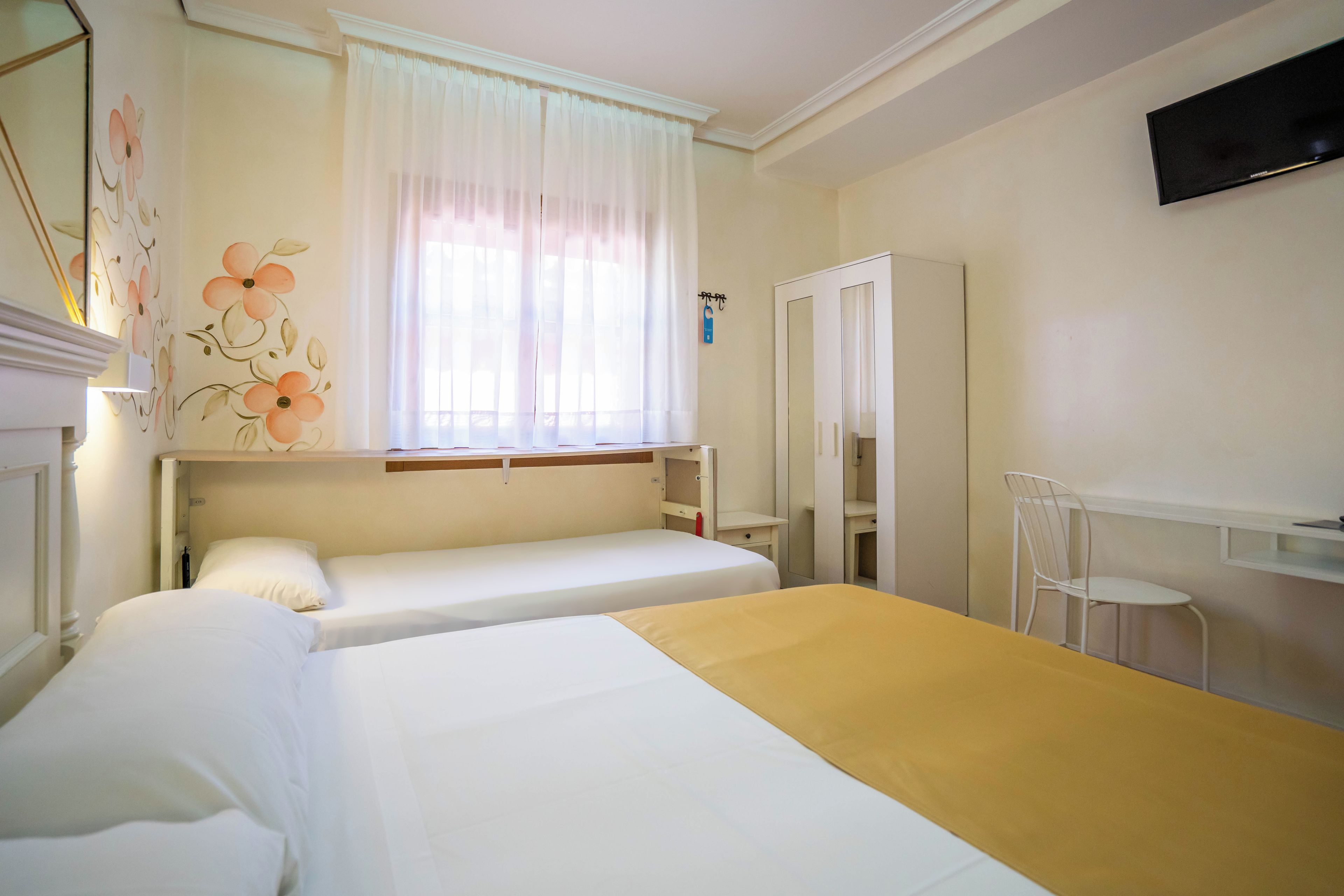 Habitació d'hotel amb dos llits individuals, una finestra amb cortines translúcides, un mirall i una petita cadira blanca.