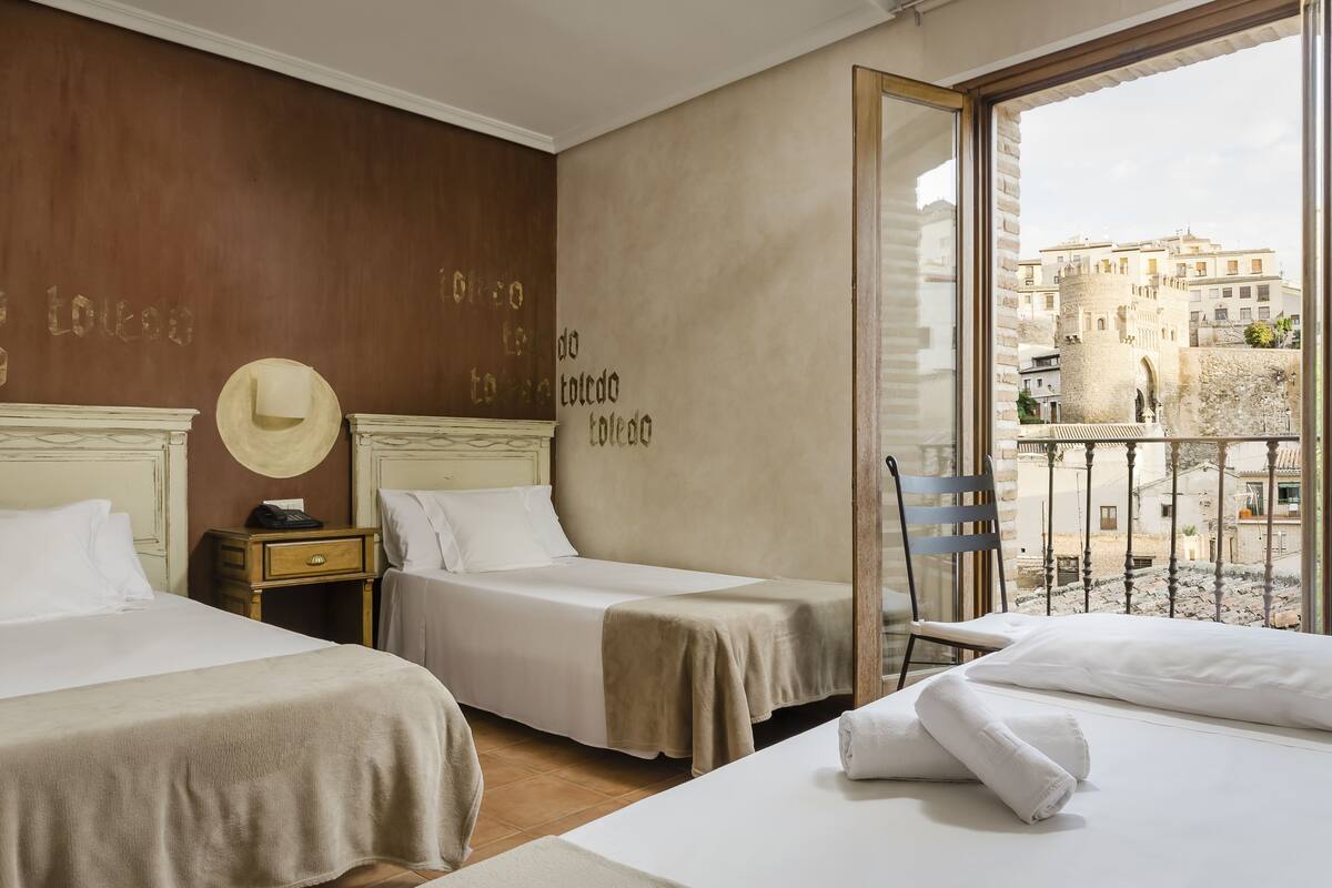 A hotel room with two beds, a wooden headboard, a nightstand, a hat on the wall, and a view of the city through a window.