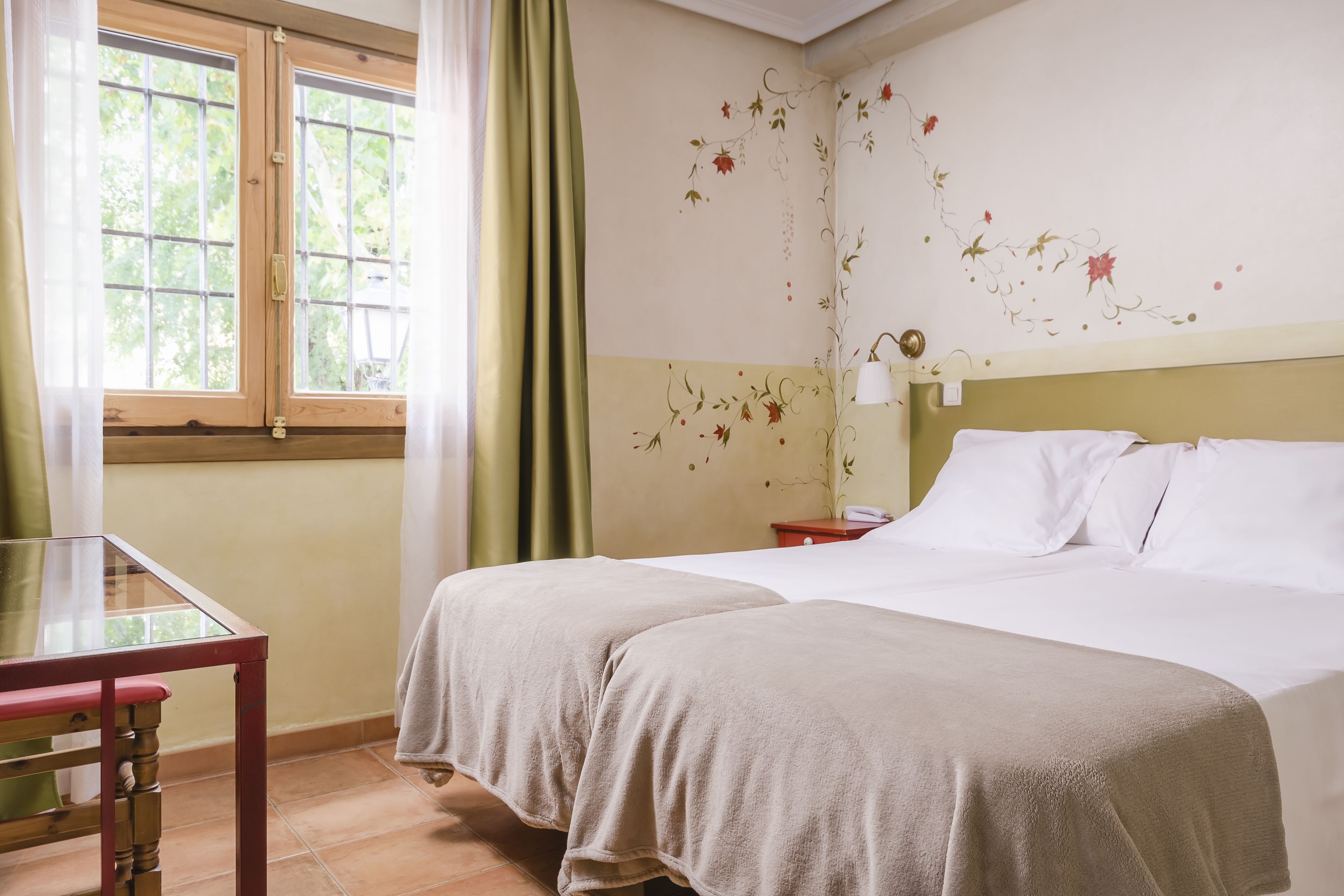 Een kamer met twee bedden, een houten tafel, een raam met gordijnen en een wand met bloemenbehang.