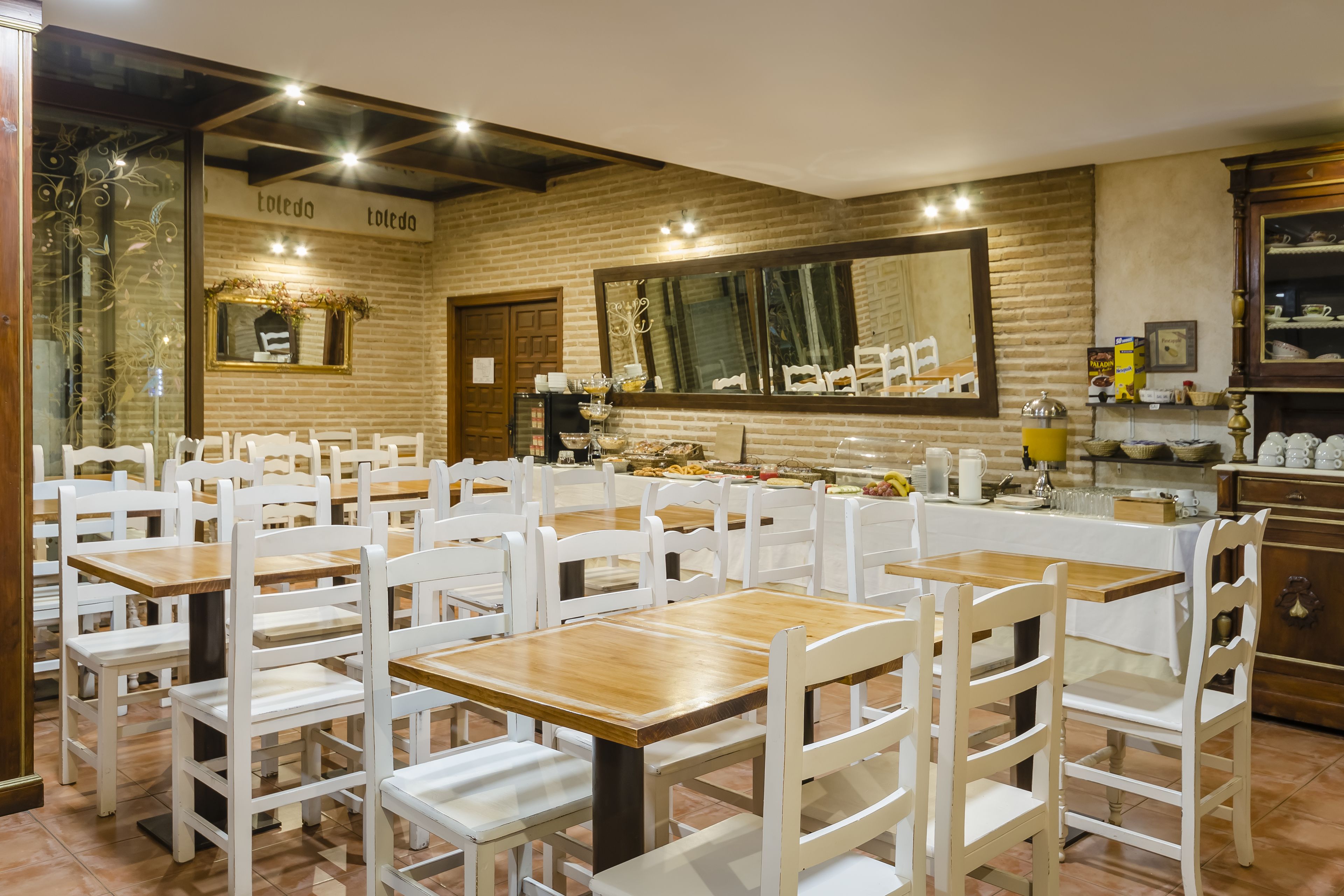 A restaurant with white chairs and wooden tables, a brick wall, and a large mirror.
