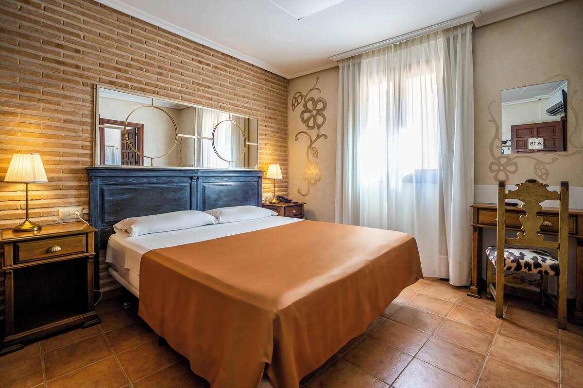 A hotel room with a brick wall, a large bed with a brown bedspread, a wooden nightstand with a lamp, a wooden dresser, and a mirror.