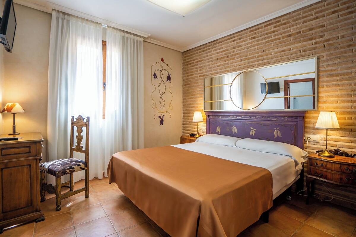 A hotel room with a large bed, a wooden chair, a desk, a dresser, and a mirror on the wall.