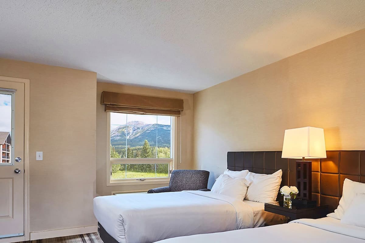 Habitación de hotel con dos camas, una mesita de noche con lámpara, ventana con vistas a la montaña y una puerta.