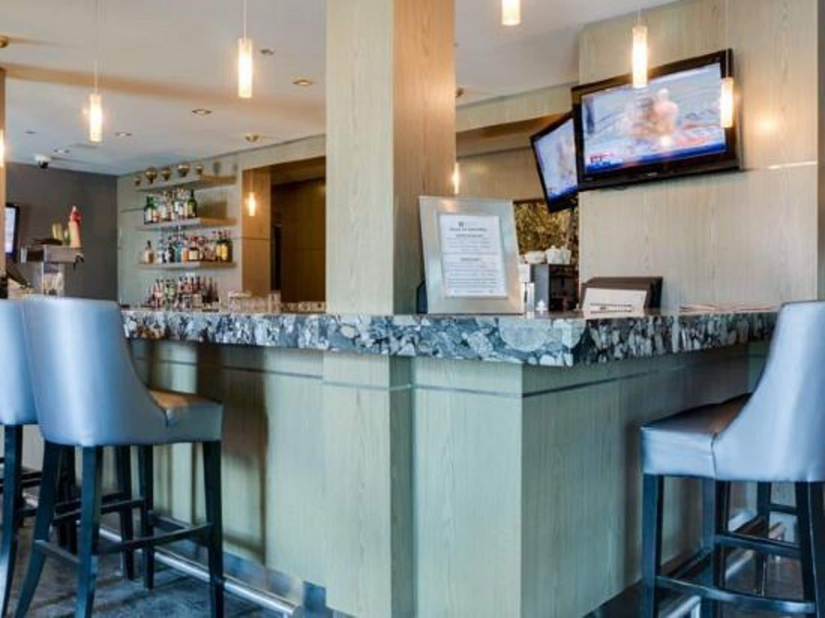 A bar area with a marble countertop, two bar stools, a television mounted on the wall, and a bar stocked with bottles.