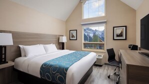 A hotel room with a large bed, a desk, a chair, a window with a mountain view, and a framed picture on the wall. - Grande Rockies Resort - Bellstar Hotels & Resorts (Canmore)