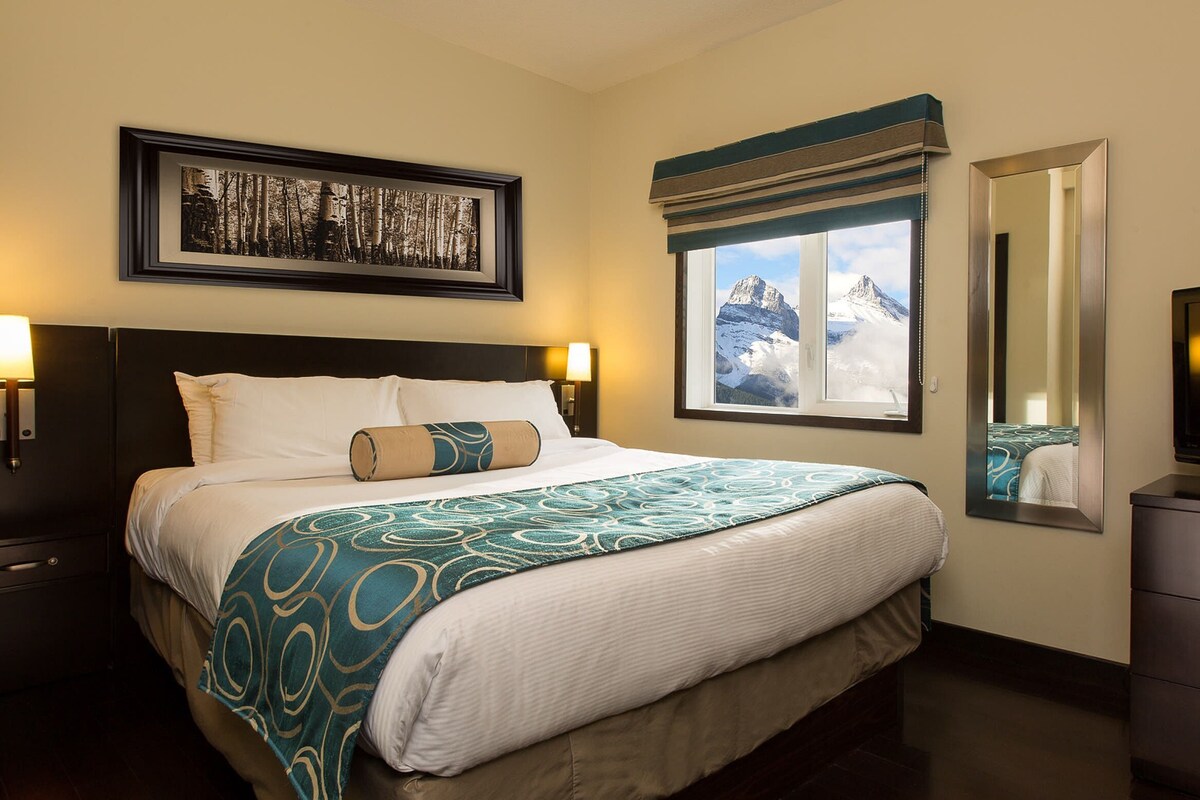 A hotel room with a bed, bedside lamps, a large window with a mountain view, a television, and a dresser.