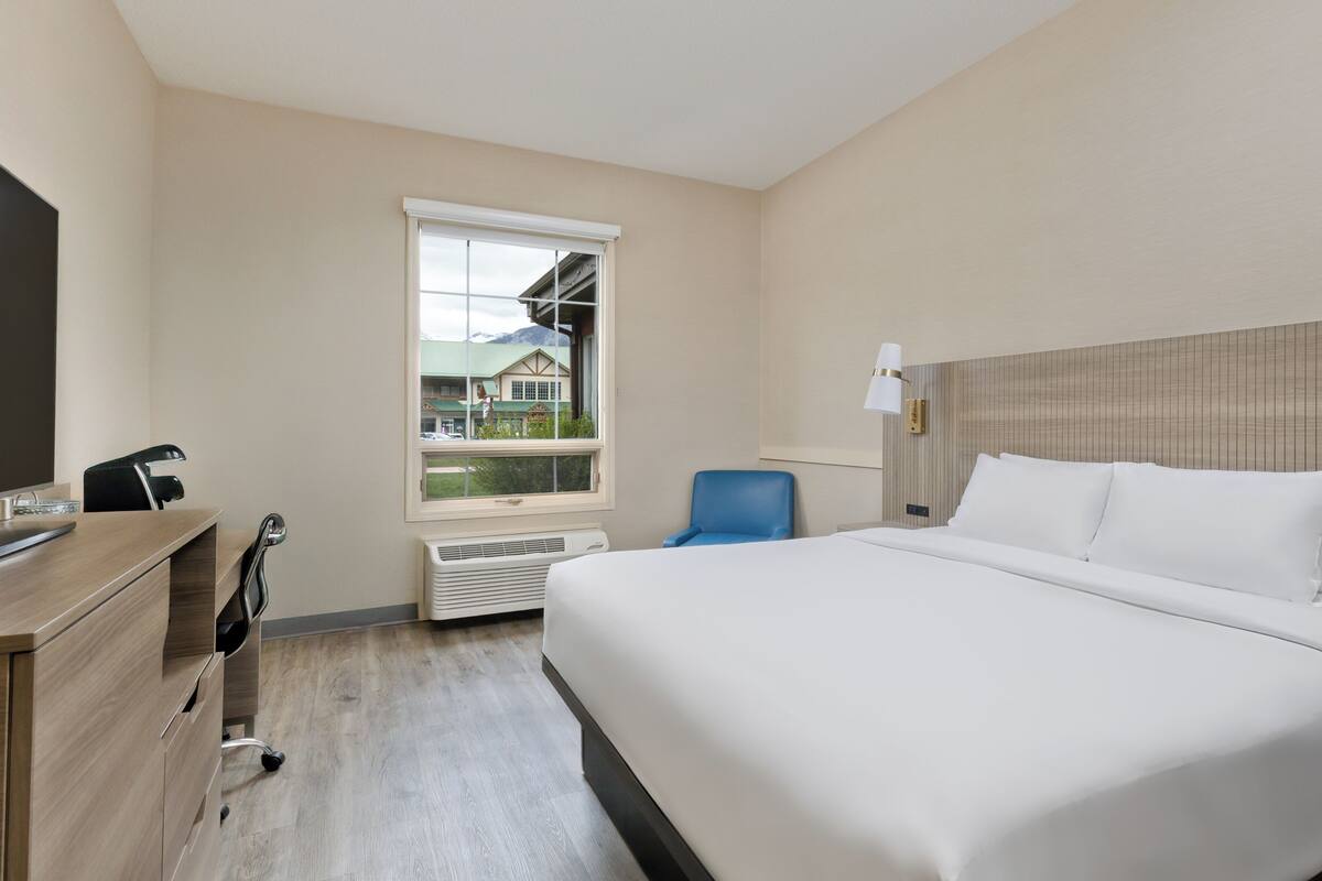 A hotel room with a bed, desk, chair, and a window with a view of buildings.