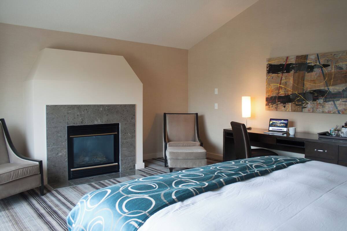 A hotel room with a fireplace, a desk with a laptop, a chair, and a bed with a patterned bedspread.