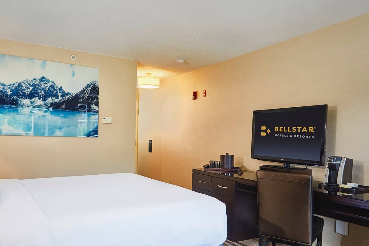 A hotel room with a bed, a desk, a television, and a painting of a mountain landscape.