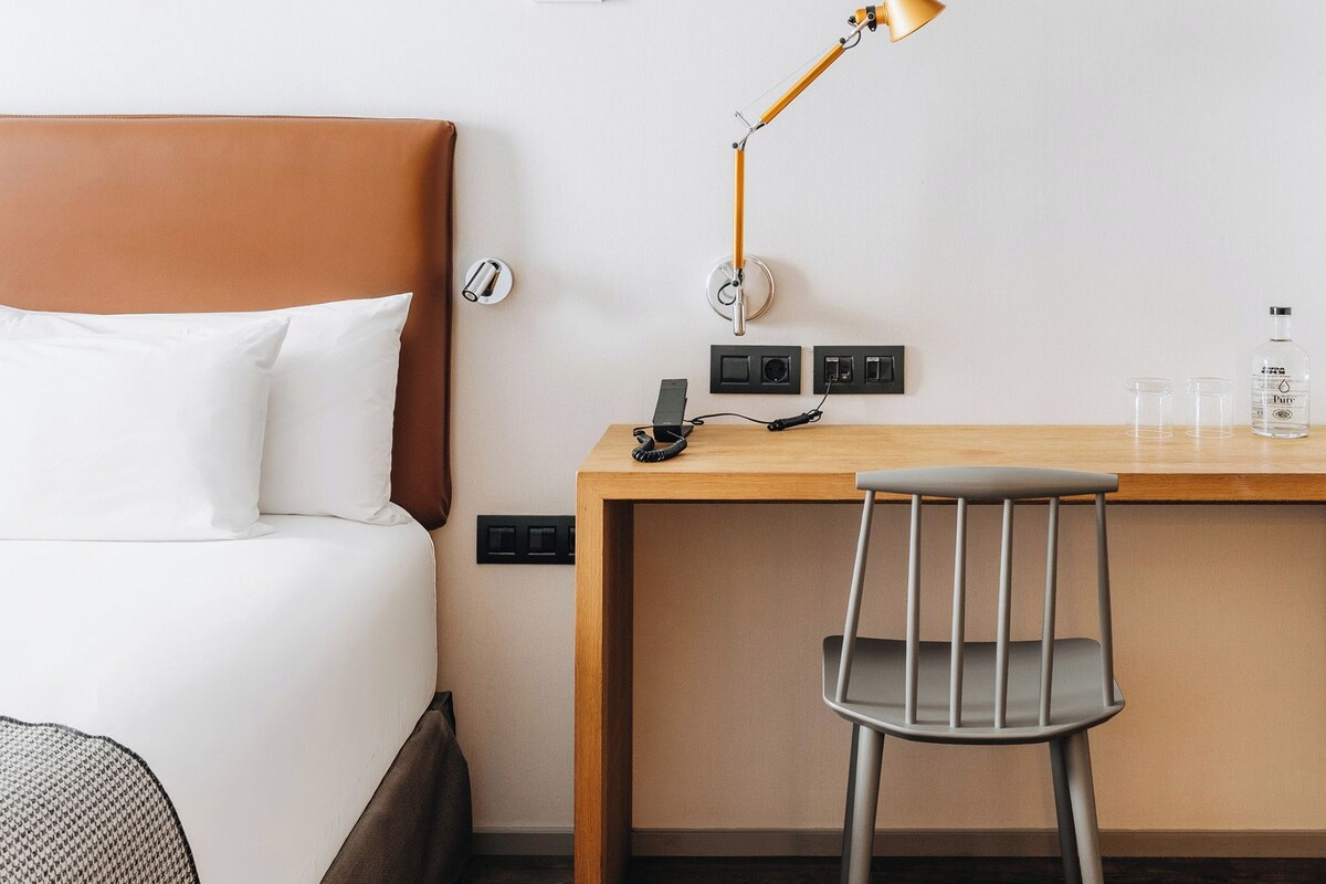 Hotelkamer met een bed, bureau, stoel, lamp, thermostaat en een waterfles.
