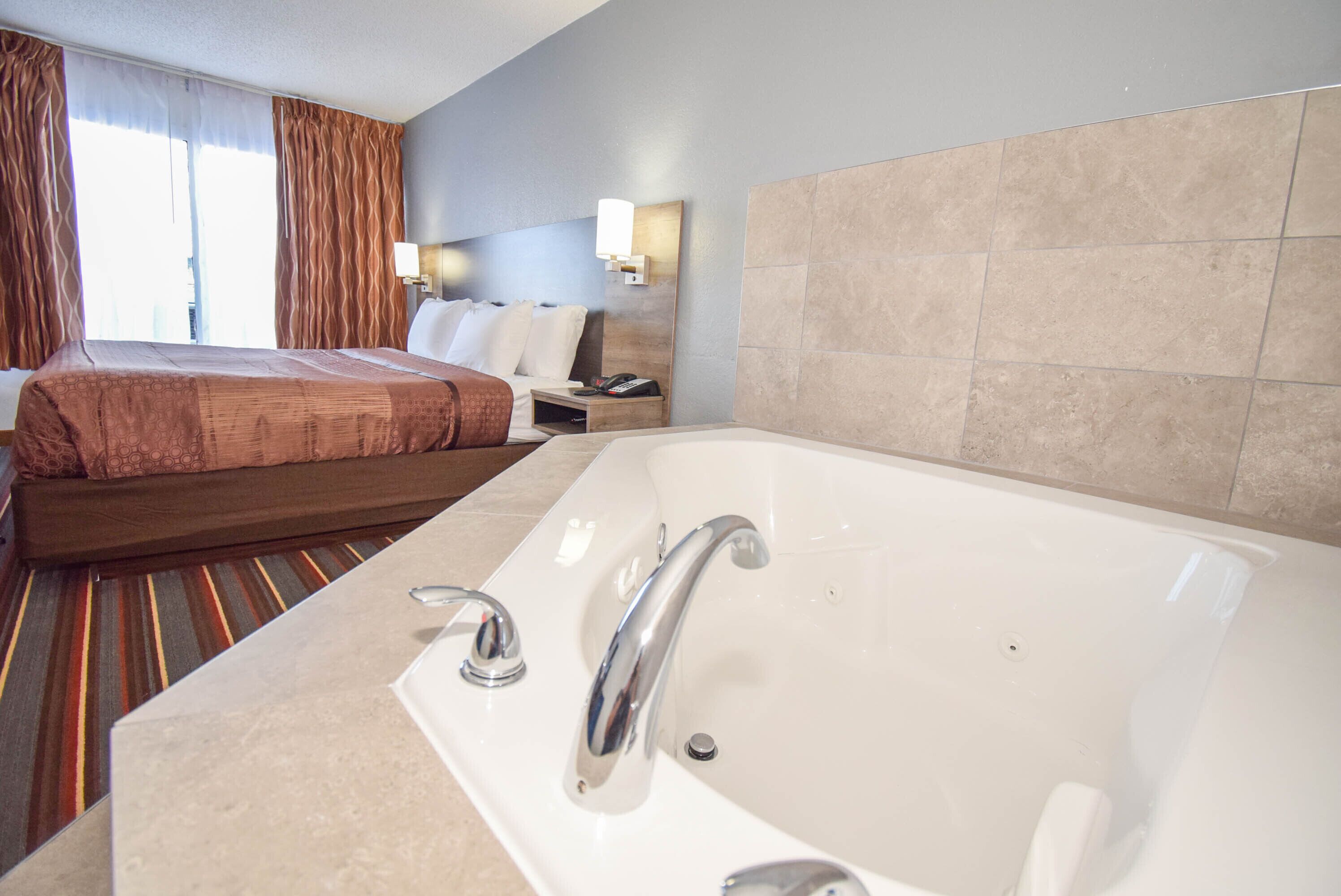 A hotel room with a large bed, a separate bathroom with a bathtub, and a nightstand with a lamp and phone.