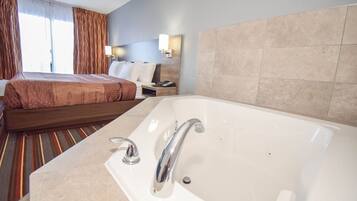 A hotel room with a large bed, a separate bathroom with a bathtub, and a nightstand with a lamp and phone.