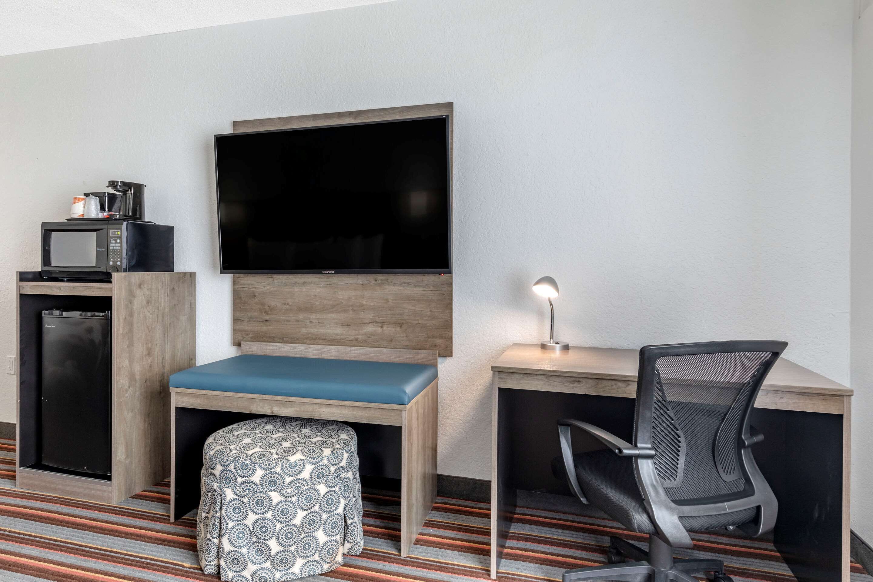 A compact hotel room with a flat-screen TV mounted on the wall, a small kitchenette with a microwave and coffee maker, a wooden desk with a chair, and a patterned ottoman.