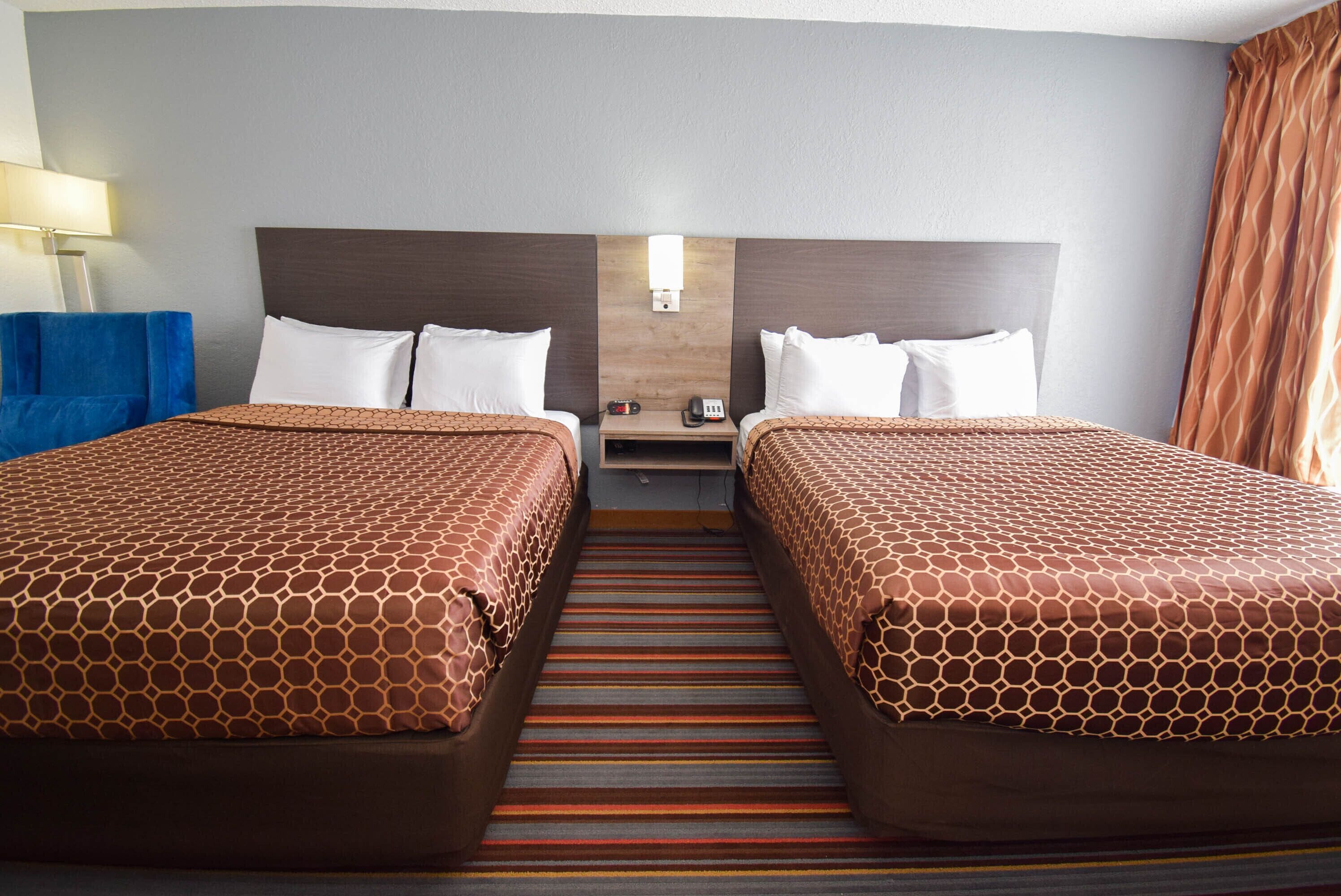 A hotel room with two beds, a nightstand, a lamp, and a view of the outside.