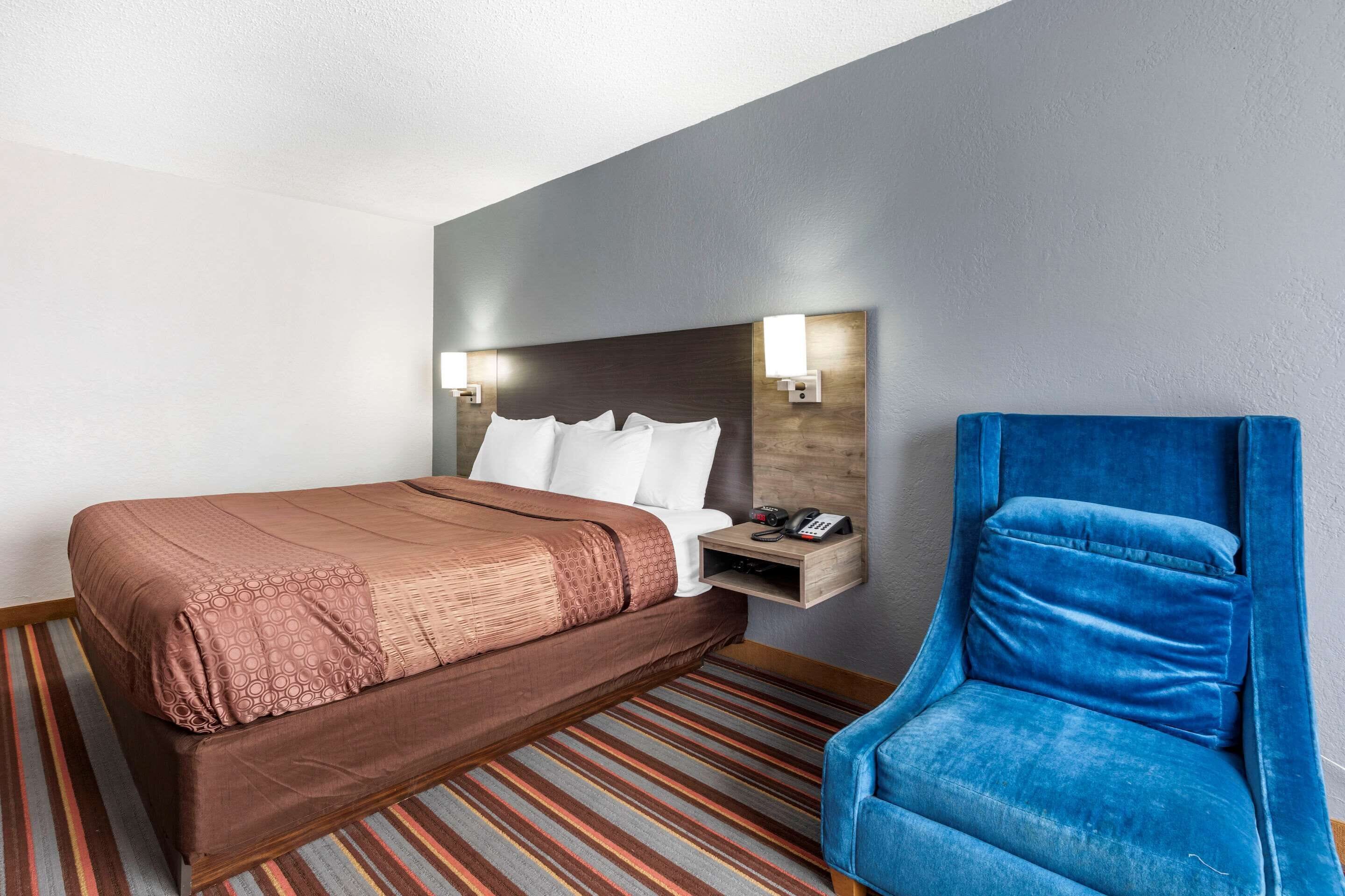 A hotel room with a bed, a blue chair, a nightstand, and a wall-mounted lamp.