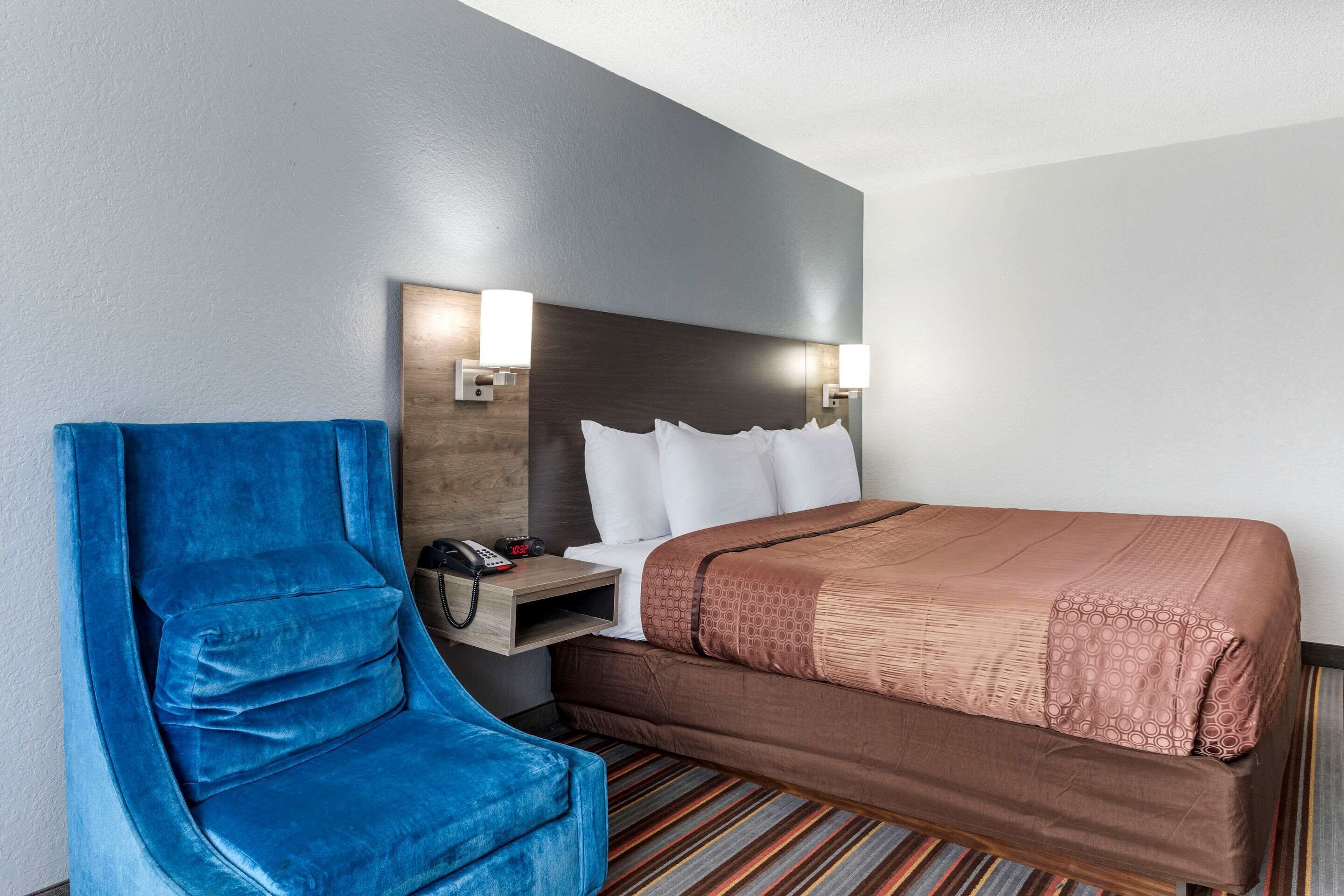 A hotel room with a large bed, a blue armchair, a bedside table with a phone, and two wall-mounted lamps.