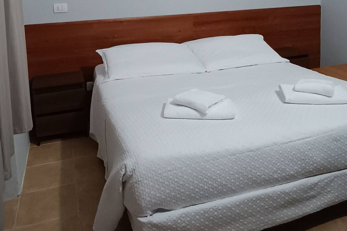A hotel room with a bed, a wooden headboard, a nightstand, and a wall socket.