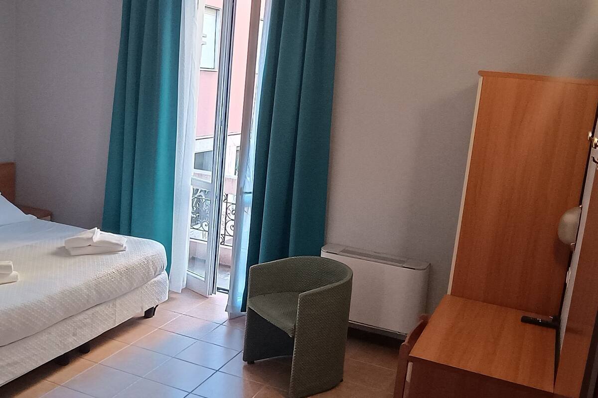 A hotel room with a bed, a chair, a wardrobe, and a window with curtains.