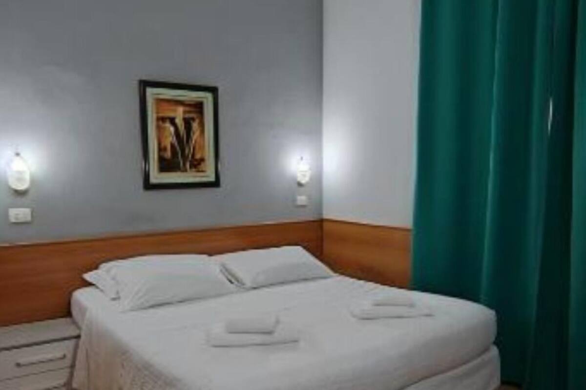 A neatly made bed in a room with a grey wall, a framed picture, and a green curtain.