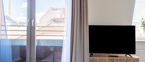 Una sala de estar moderna con un televisor grande sobre un mueble de madera, un ventanal corredero que da a un balcón y una planta en maceta en el alféizar.