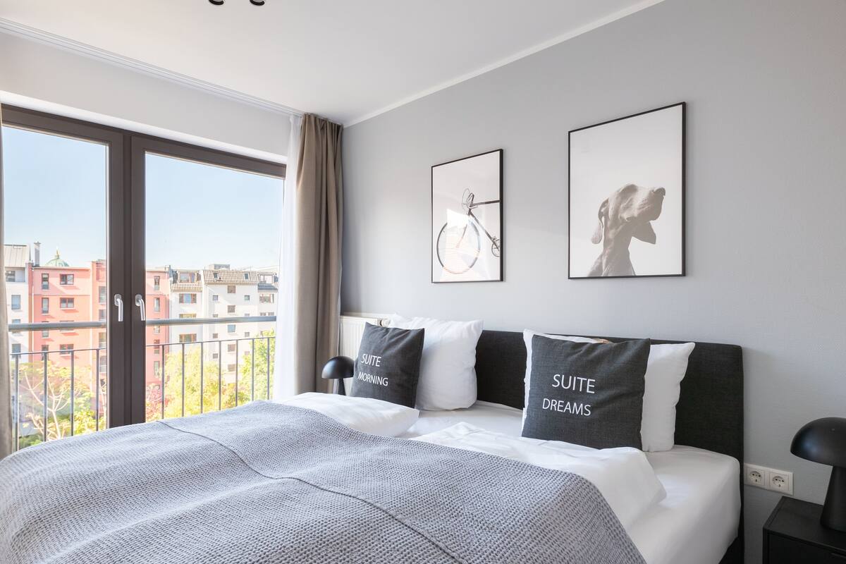 Habitación de hotel moderna con una cama grande, dos almohadas con texto, vista a edificios y un cuadro enmarcado en la pared.