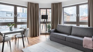 Una sala moderna con un sofá gris, una mesa de comedor con sillas, una lámpara de pie y ventanas grandes con cortinas.