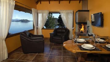 Un área de comedor con vistas a un lago y montañas, sillones de cuero, chimenea y una mesa de comedor preparada para cuatro.