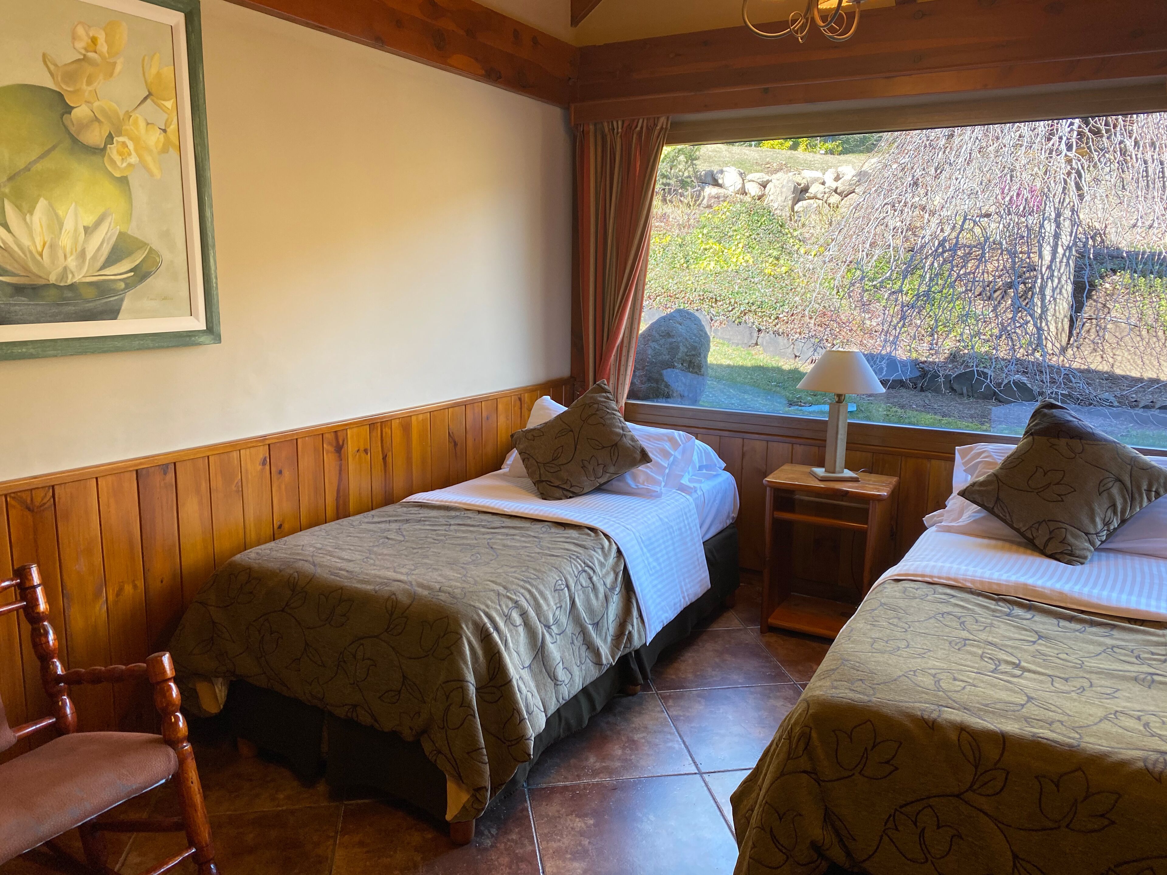 A room with two beds, wooden walls, a window with a view of greenery, a painting on the wall, and a lamp on a bedside table.