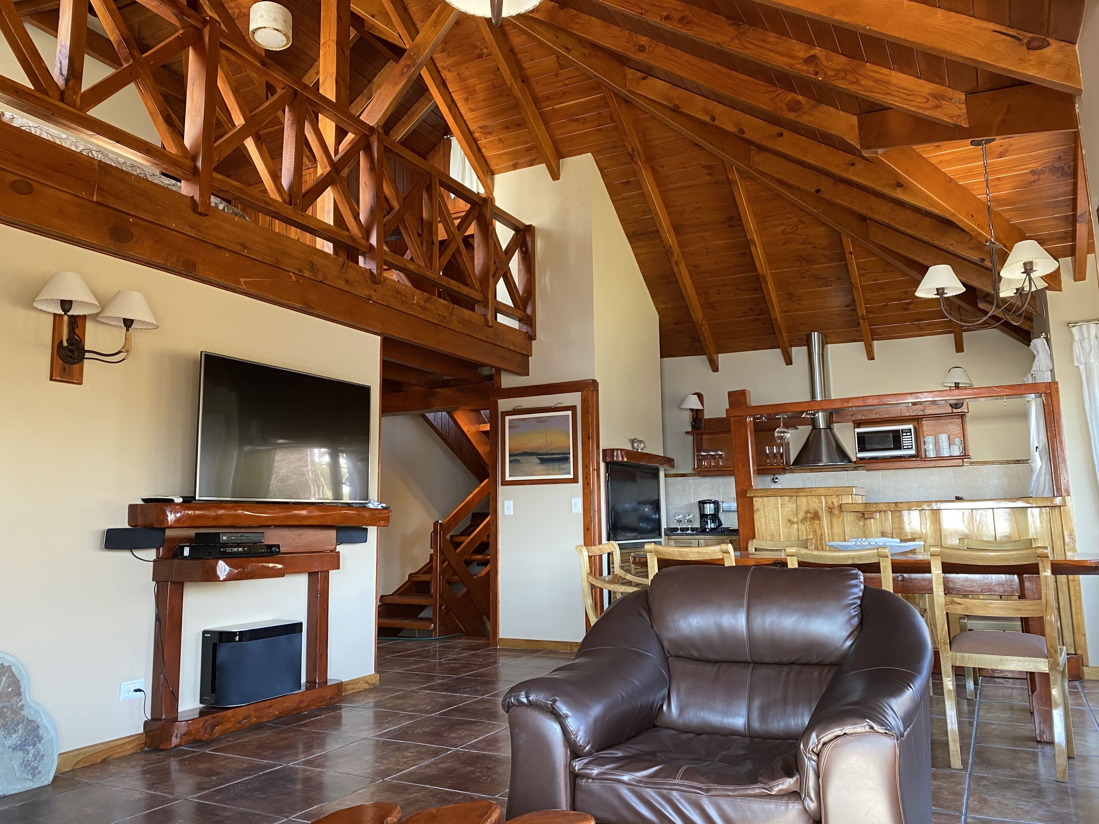 A spacious living area with a high ceiling, wooden beams, a fireplace, a leather sofa, and a kitchen area with a microwave and coffee maker.