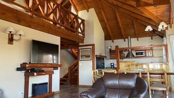 A spacious living area with a high ceiling, wooden beams, a fireplace, a leather sofa, and a kitchen area with a microwave and coffee maker.